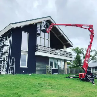 Rode hoogwerker met de bak bij de dakrand van een donker afgewerkte woning met balkon.