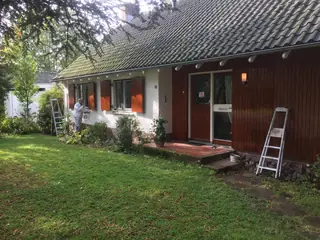 Twee ladders tegen een langgerekte woning met houten gevelbekleding, met een schilder aan het werk.