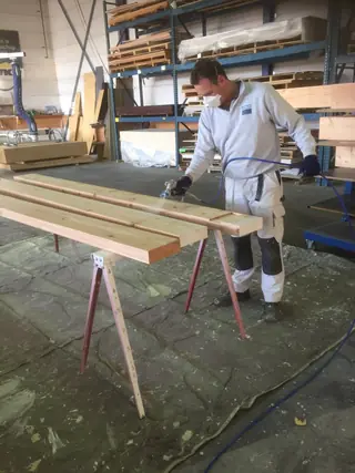 Schilder spuit houten balken op schragen in een werkplaats.