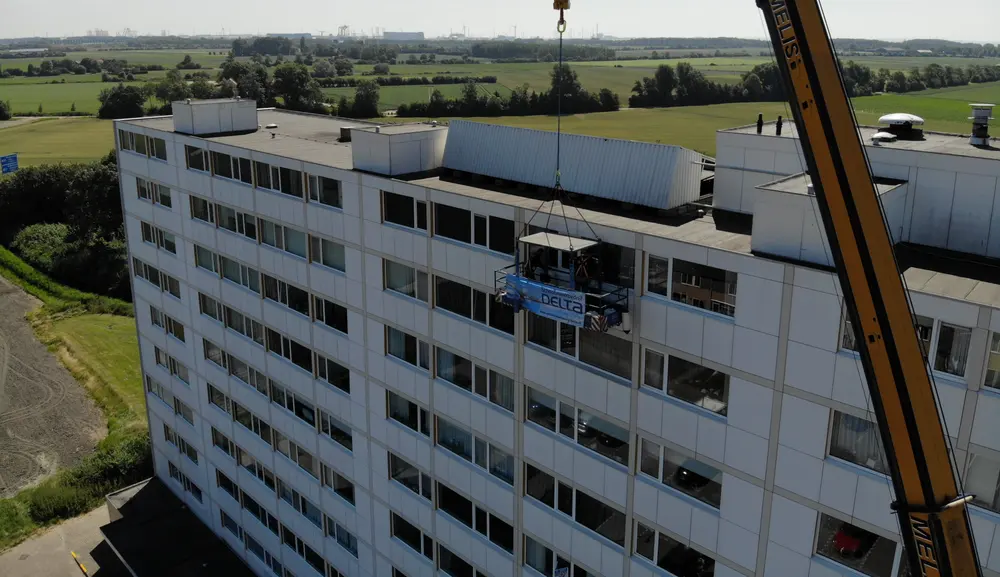 Galerijflat op Walcheren, gezien vanuit de lucht, met een werkbak van Delta halverwege de gevel aan een gele torenkraan en de polder erachter.