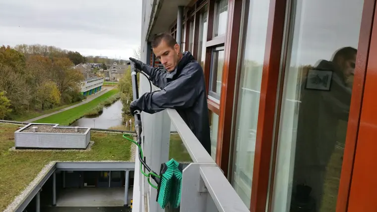 Medewerker van Delta zeemt een raam terwijl hij over een balustrade leunt, met een kanaal en weiland achter zich.