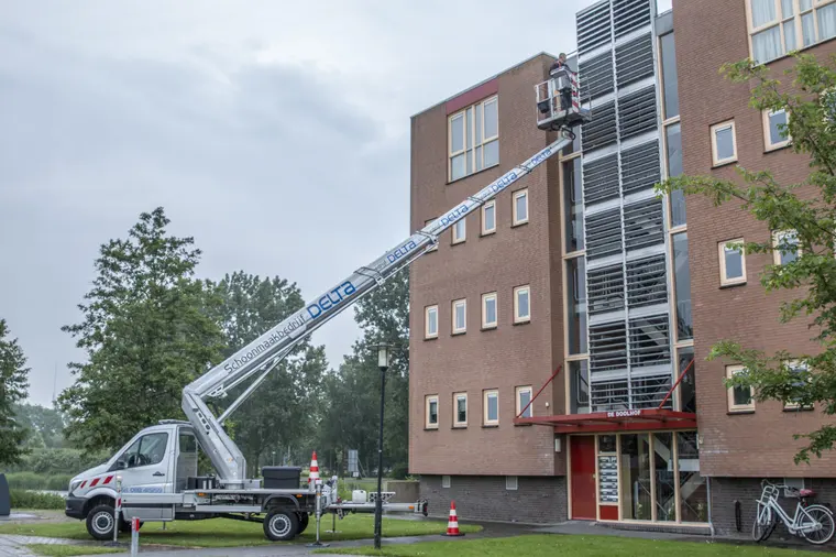 De eigen hoogwerker van Delta staat met de giek omhoog naast een bakstenen flat, met pylonen op straat ervoor.