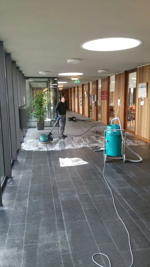 Lange gang met een donkere tegelvloer waarin een medewerker van Delta met een schrobmachine werkt.