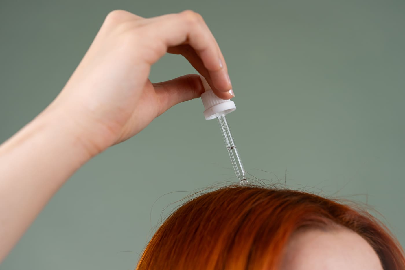 Een hand druppelt met een pipet serum op de hoofdhuid tussen roodbruin haar.