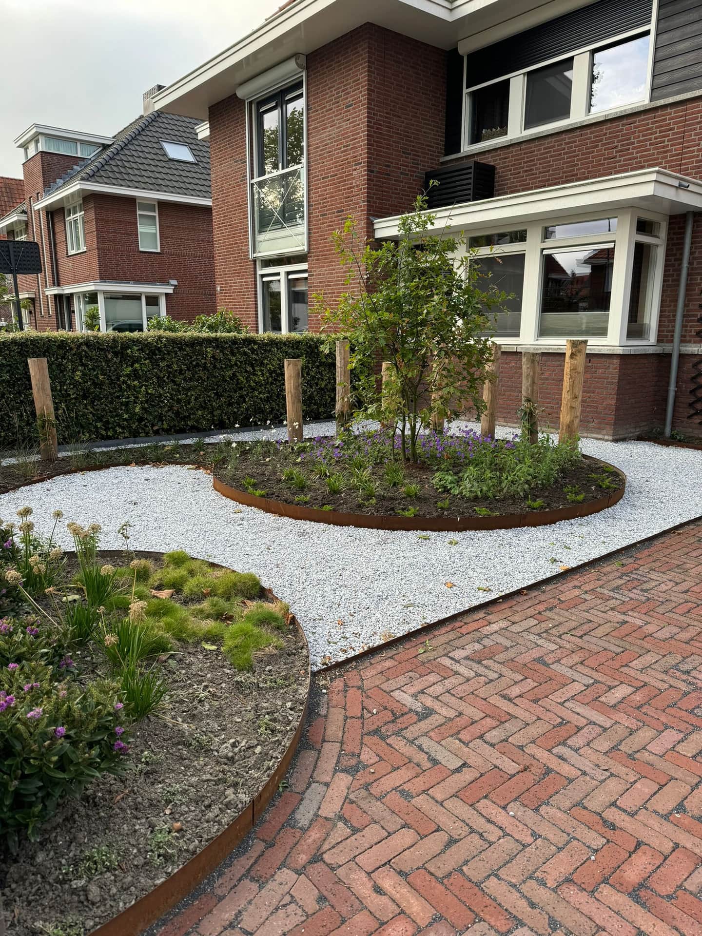 Voortuin met wit split, cortenstalen randen, jonge beplanting en een pad van rode klinkers