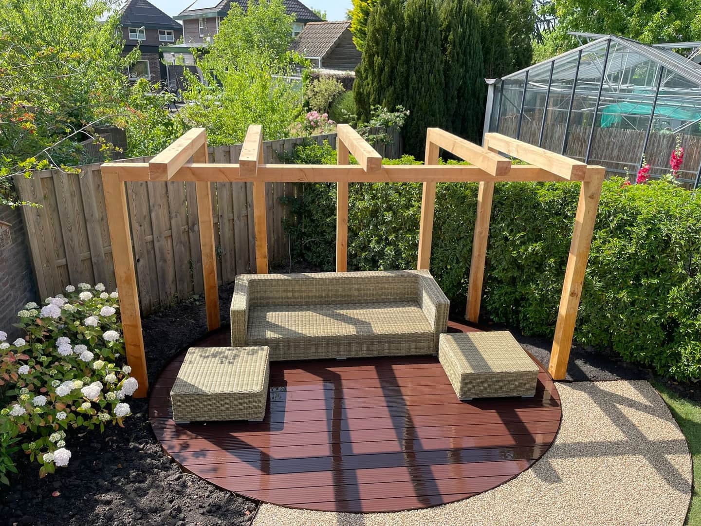 Houten pergola boven een rond hardhouten terras met loungeset en volgroeide haag