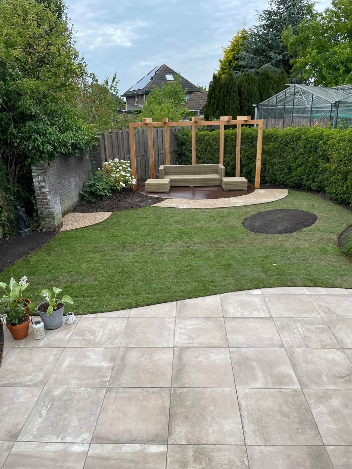 Achtertuin met ruim gazon, groot tegelterras en een houten pergola bij de haag