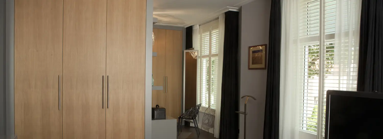 Slaapkamer met een kastenwand in licht eiken en een doorkijk naar een kleedruimte.