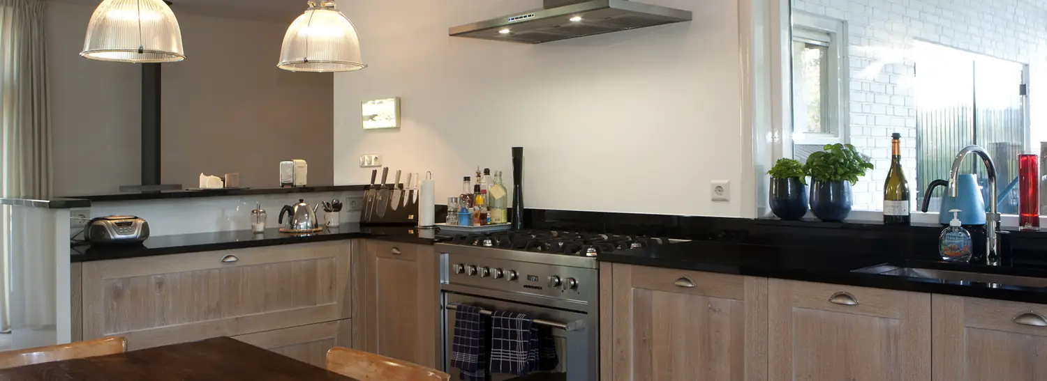 Keuken in licht eiken met een donker werkblad, een roestvrijstalen fornuis en twee hanglampen boven de eettafel.