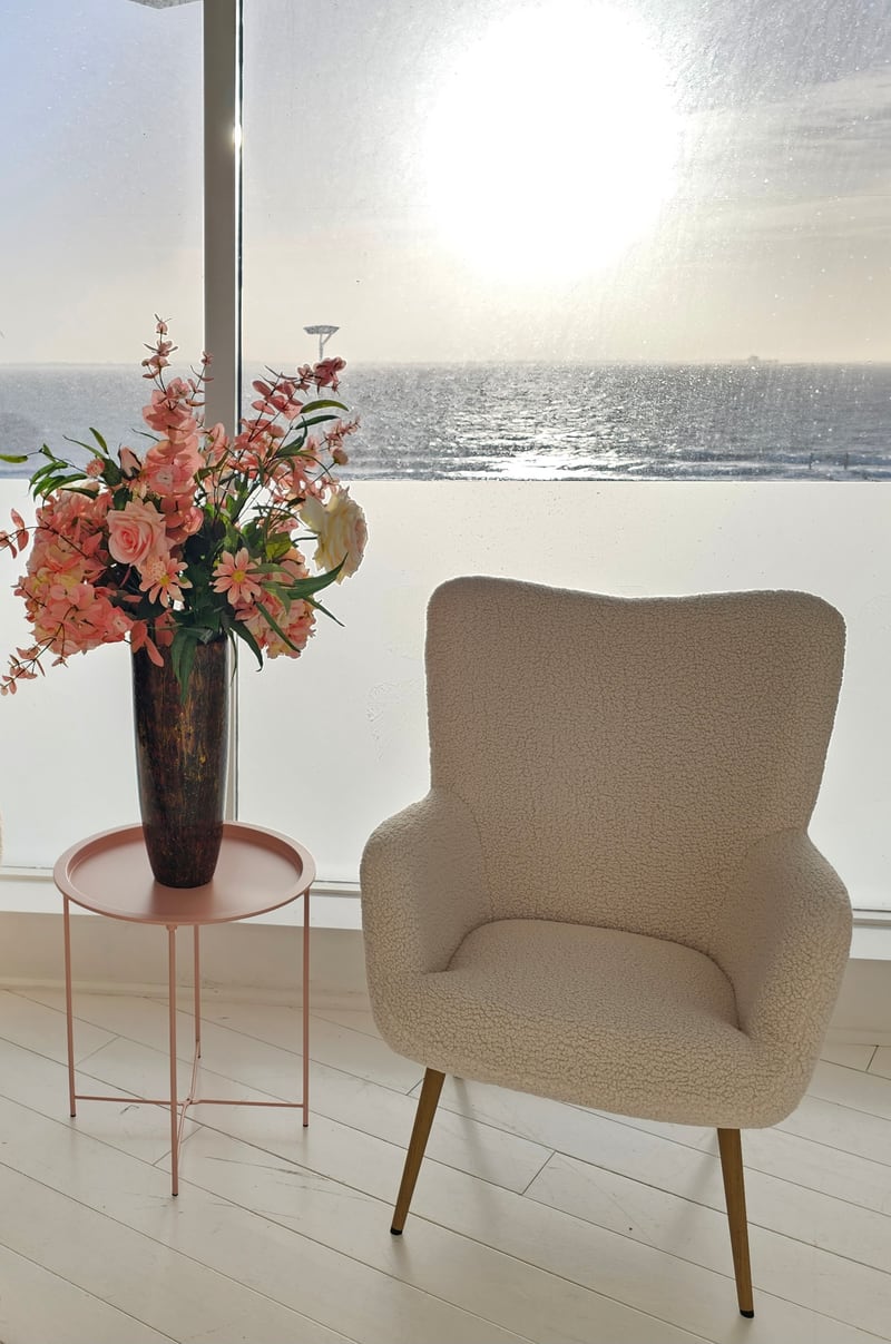 Een cr&egrave;mekleurige boucl&eacute;stoel en een oud roze bijzettafel met roze bloemen voor een raam waarachter de zon boven zee staat.