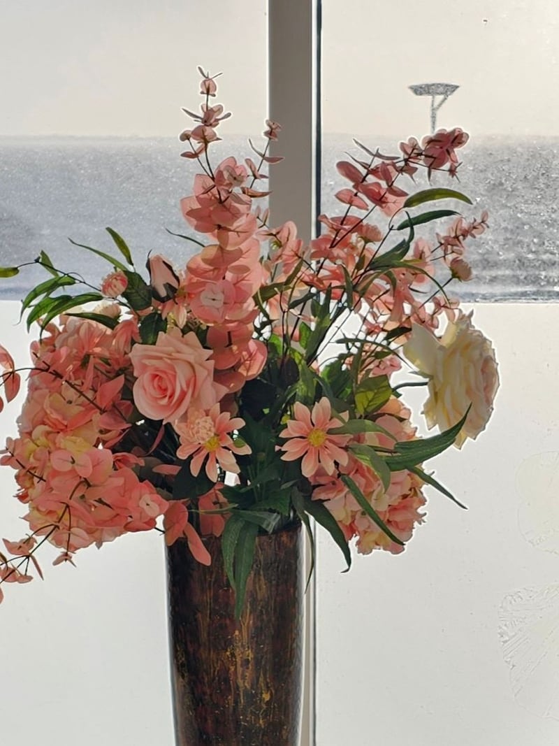 Een boeket roze bloemen in een donkere vaas voor het raam van de salon, met de zee erachter.