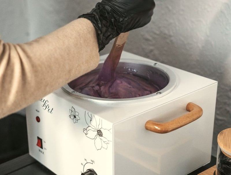 Een gehandschoende hand roert met een houten spatel in een pot met gesmolten wax.
